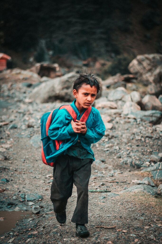 PROVISION OF NECESSITIES FOR CHILDREN A young child in a blue jacket walks with a backpack through rocky terrain in Kel, showcasing determination.