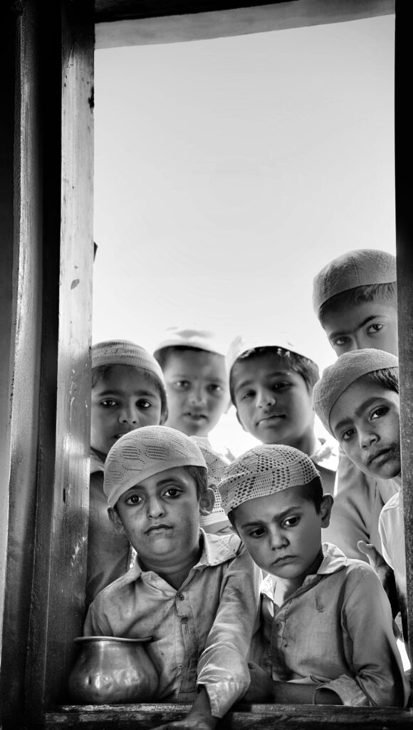 WE EDUCATE A group of children in traditional attire gazing through a window with solemn expressions.